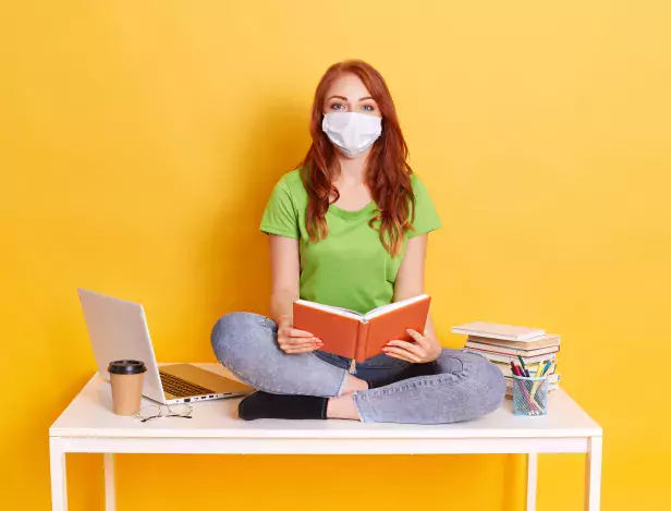 young-student-in-medical-mask-studying-at-home-while-quarantine-bored-of-distance-learning-sitting-with-crossed-legs-on-white-table-with-book-in-hands